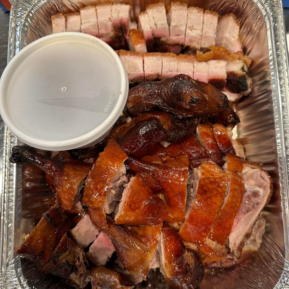 A foil tray filled with sliced roasted duck, crispy pork belly, and a white sauce container.